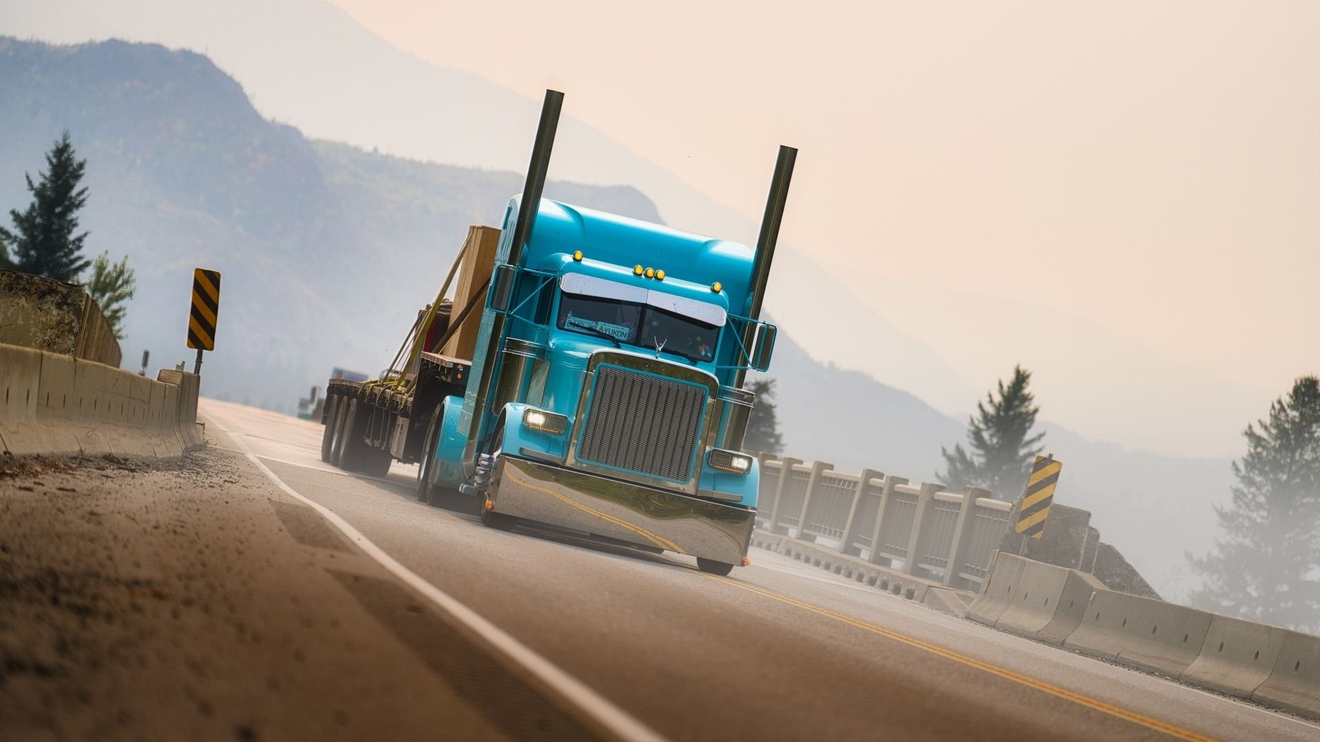 Blue semi-truck driving on mountain highway with protective barriers and forested landscape